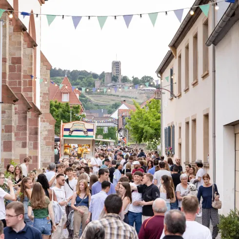 Eine lebhafte Straße voller Menschen mit bunten Wimpeln im Hintergrund. Im Hintergrund sind Gebäude und eine Hügelkulisse zu sehen.