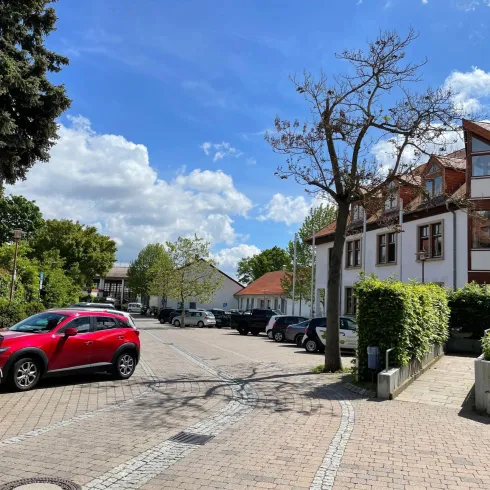 Eine ruhige Straße mit parkenden Autos und Bäumen. Im Hintergrund sind Häuser und ein blauer Himmel mit einigen Wolken zu sehen.