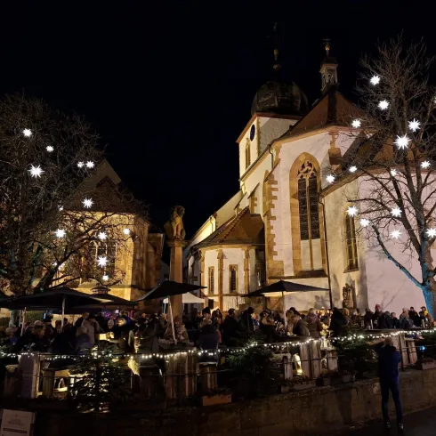 Ein belebter Platz bei Nacht mit festlicher Beleuchtung und einer Kirche im Hintergrund. Viele Menschen genießen die Atmosphäre unter den leuchtenden Sternen.