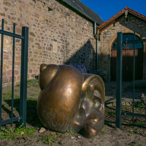 Eine bronzene Skulptur vor einer alten Steinmauer. Im Hintergrund ist ein historisches Gebäude mit einem roten Dach zu sehen.