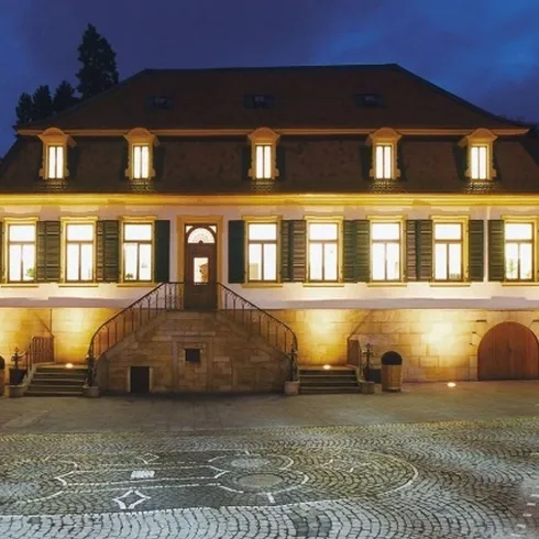 Ein historisches Gebäude bei Nacht, beleuchtet von warmem Licht. Der Platz vor dem Gebäude ist mit Steinen gepflastert.