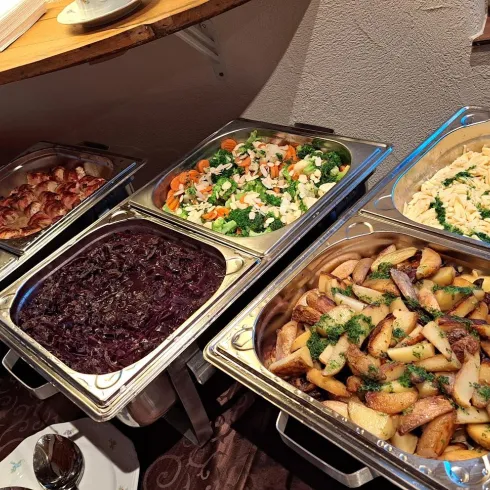Ein Buffet mit verschiedenen warmen Speisen, darunter Fleisch, Salat, Kartoffeln und Pasta. Die Gerichte sind appetitlich angerichtet und bereit zum Servieren.