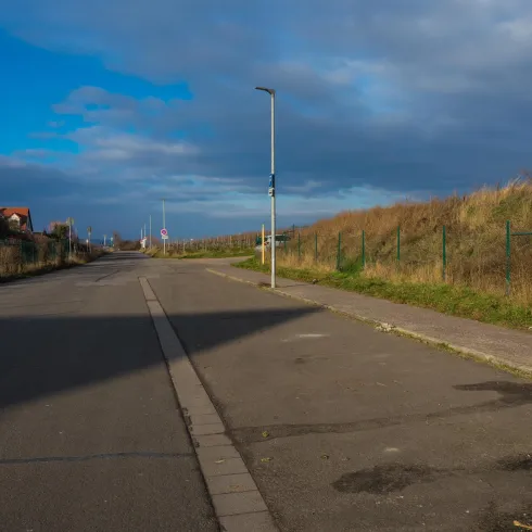 Eine verlassene Straße mit einem Bürgersteig und Grasbewuchs an den Seiten. Der Himmel ist teilweise bewölkt und die Umgebung wirkt ruhig.
