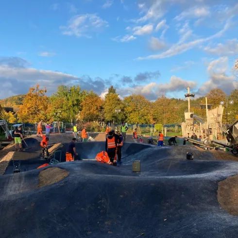 Eine Baustelle mit vielen Arbeitern, die an einem Gelände für Sportaktivitäten arbeiten. Im Hintergrund sind Bäume mit buntem Herbstlaub und ein Baugerät zu sehen.