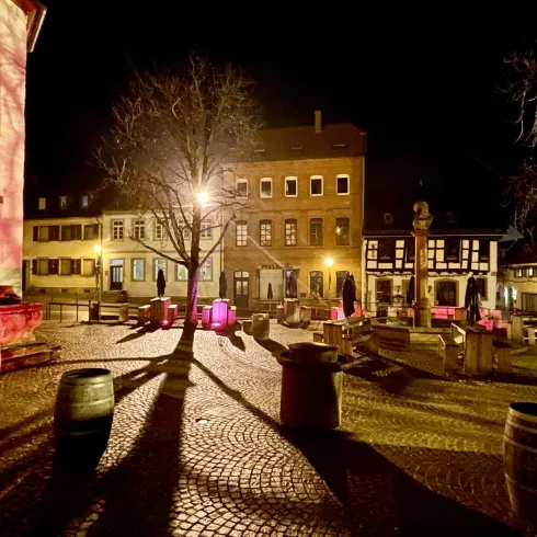 Ein ruhiger Platz bei Nacht mit warmem Licht und Schatten von Bäumen. Die Umgebung ist charmant und einladend, mit historischen Gebäuden und Sitzgelegenheiten.