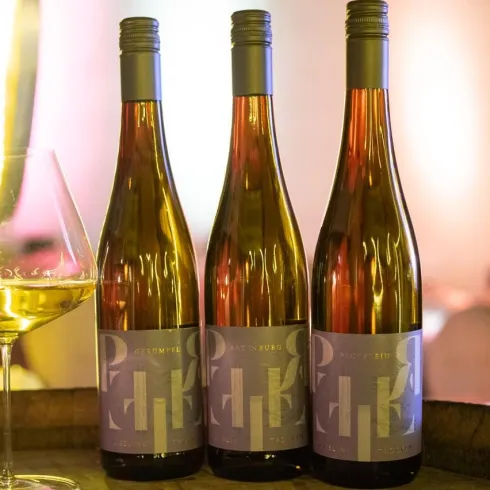 Drei Flaschen Wein stehen auf einem Holzfass, daneben ein Glas mit Weißwein. Die Umgebung ist warm beleuchtet und schafft eine einladende Atmosphäre.