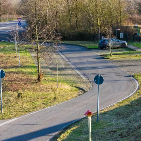 Eine Kurve auf einer Straße mit Verkehrsschildern und Parkplatz. Im Hintergrund sind Bäume und eine grüne Landschaft zu sehen.