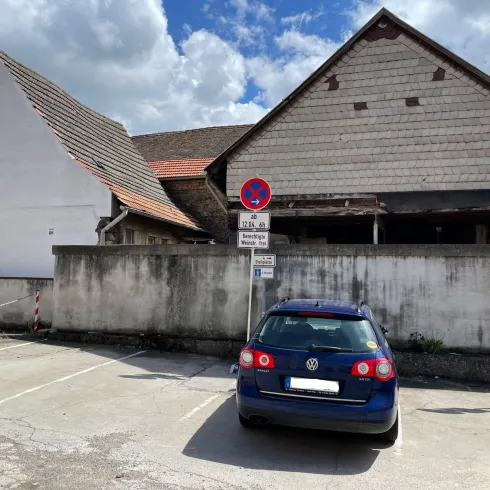 Ein blauer Volkswagen parkt vor einem alten, teils maroden Gebäude. Im Hintergrund sind Verkehrssignale und ein bewölkter Himmel zu sehen.
