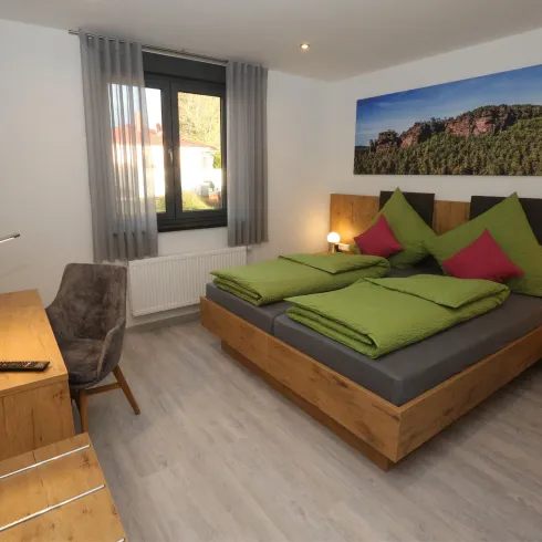 Ein modernes Schlafzimmer mit einem Doppelbett und grünen Kissen. Der Raum ist hell und gemütlich, mit einem Schreibtisch und einem großen Fenster.