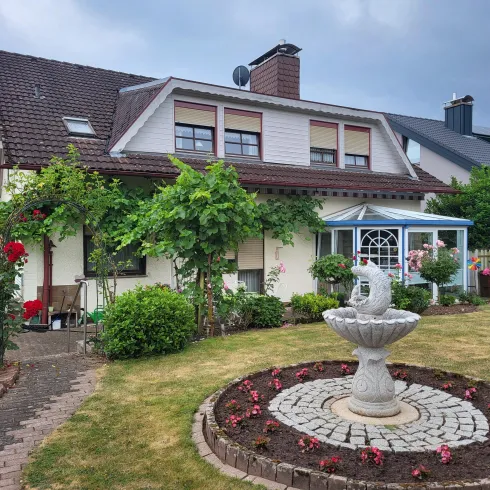 Ein Einfamilienhaus mit einem schön gestalteten Garten. Im Vordergrund steht ein Brunnen, umgeben von Blumen und Sträuchern.
