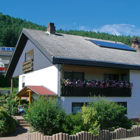 Ein modernes Einfamilienhaus mit einem Balkon und einem gepflegten Garten. Im Hintergrund sind bewaldete Hügel zu sehen.