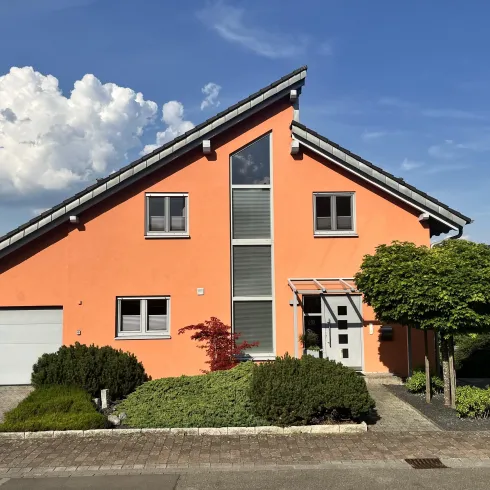 Ein modernes, orangefarbenes Haus mit einem schrägen Dach und großen Fenstern. Die Umgebung ist gepflegt mit grünen Pflanzen und einem Parkplatz.