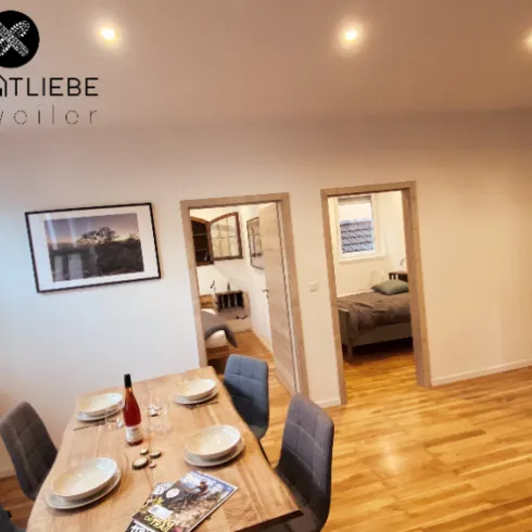 Ein stilvolles Wohnzimmer mit einem großen Esstisch und modernen Stühlen. Im Hintergrund sind ein Schlafzimmer und ein Badezimmer sichtbar.