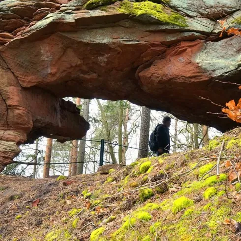 Eine natürliche Felsformation mit einem großen Durchgang und grünem Moos. Im Hintergrund ist eine Person zu sehen, die den Weg entlang läuft.