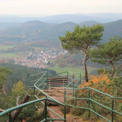 Ein Aussichtspunkt mit einem Blick auf ein malerisches Dorf inmitten von Hügeln. Bunte Herbstblätter umgeben den Weg zum Aussichtspunkt.