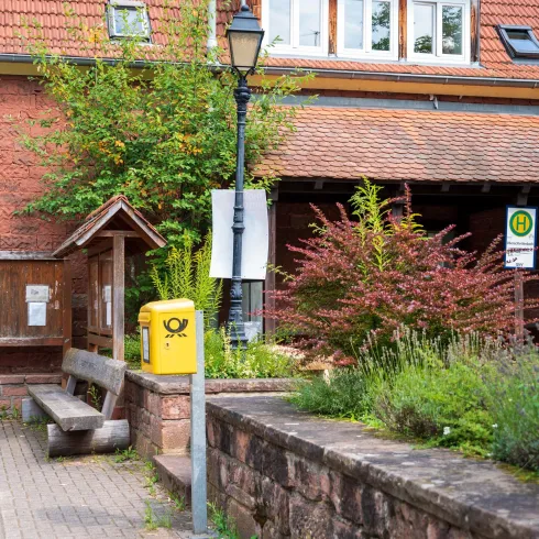 Ein gemütlicher Ort mit einem Holzbank und einem gelben Briefkasten. Die Umgebung ist mit Bäumen und Büschen begrünt.