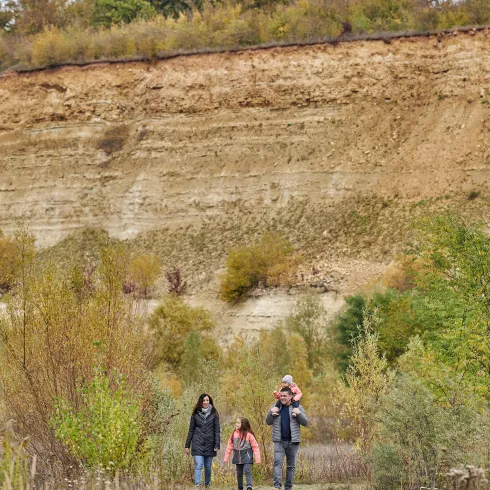 Eine Familie wandert in einer naturnahen Landschaft. Im Hintergrund sind steile Erdwände und farbenfrohes Laub zu sehen.