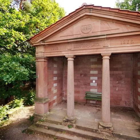 Ein historischer Pavillon mit Säulen steht in einer grünen Umgebung. Die Fassade besteht aus rotem Stein und zeigt das Jahr 1896.