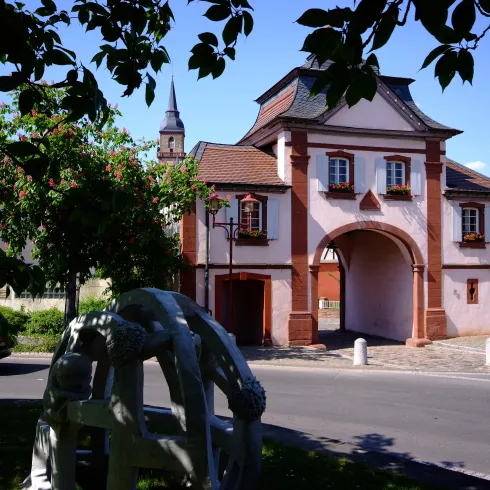 Ein historisches Gebäude mit einem charakteristischen Tor in einer kleinen Stadt. Im Vordergrund sind Bäume und eine Skulptur sichtbar.