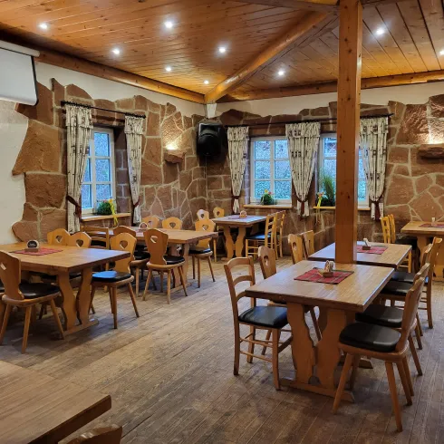 Ein einladendes Restaurant mit Holztischen und Stühlen. Die Wände sind aus Stein, und große Fenster lassen viel Licht herein.
