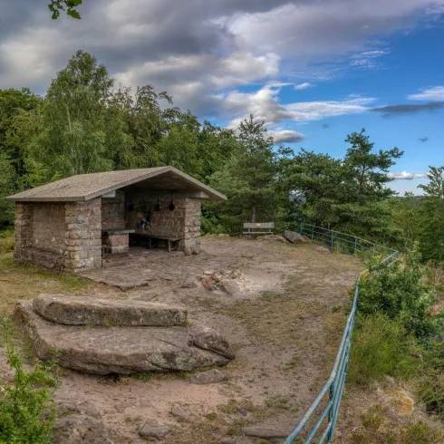 Eine kleine Steinhütte auf einem Felsen, umgeben von Bäumen und einem weiten Himmel. Der Ort bietet eine ruhige Aussicht auf die Natur.