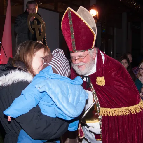 Eine festliche Szene mit einem freundlichen Mann im Gewand des Nikolaus, der ein Kind umarmt. Im Hintergrund sind viele Menschen zu sehen, die die Veranstaltung beobachten.