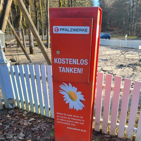 Eine rote Ladesäule für E-Bikes mit der Aufschrift „KOSTENLOS TANKEN!“ steht vor einem Biergarten. Im Hintergrund sind Spielgeräte und ein eingezäunter Bereich zu sehen.