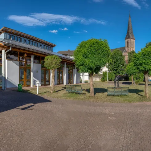 Ein ruhiger Platz mit modernen Gebäuden und runden Bäumen. Im Hintergrund ist eine Kirche mit einem hohen Turm sichtbar.