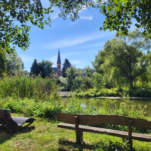 Ein ruhiger Park mit zwei Bänken am Wasser. Im Hintergrund ist ein Kirchturm und viel grüne Landschaft zu sehen.