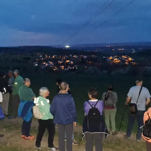 Eine Gruppe von Menschen steht auf einem Hügel und schaut auf ein beleuchtetes Dorf im Abendlicht. Der Himmel ist leicht bewölkt und es ist spät am Tag.