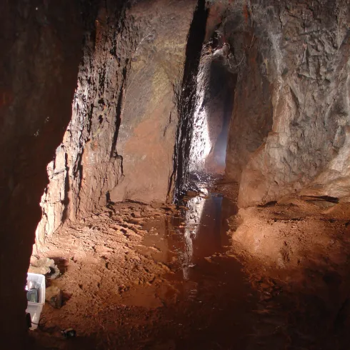 Eine dunkle Höhle mit feuchten Wänden und sichtbar mineralischen Ablagerungen. Lichtstrahlen fallen in den Raum und reflektieren auf dem Boden.