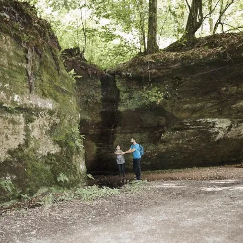 Eine beeindruckende Felsformation in einem Wald. Zwei Personen stehen auf einem Weg und erkunden die Umgebung.