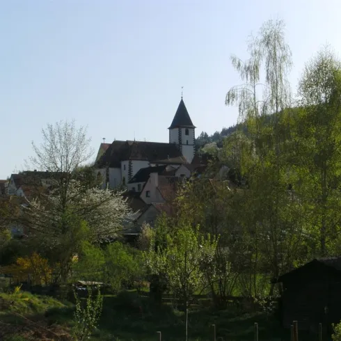 Eine malerische Landschaft mit einem kleinen Dorf und einer Kirche im Hintergrund. Umgeben von Bäumen und blühenden Pflanzen strahlt die Szene Ruhe und Idylle aus.