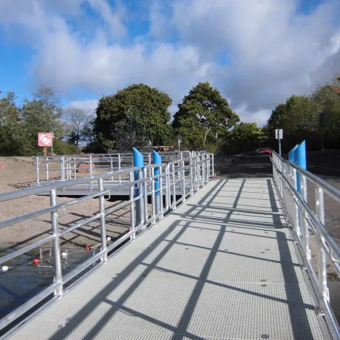 Eine moderne Rampe mit Geländer führt zum Wasser. Im Hintergrund sind Bäume und ein bewölkter Himmel zu sehen.