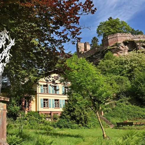 Eine malerische Landschaft mit einem historischen Gebäude und einer Statue im Vordergrund. Im Hintergrund sind Bäume und eine Burgruine sichtbar.