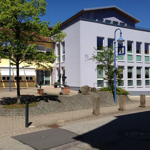 Ein modernes, lila Gebäude mit großen Fenstern steht an einem ruhigen Platz. Umgeben von Bäumen und einer gepflasterten Fläche, strahlt es eine angenehme Atmosphäre aus.