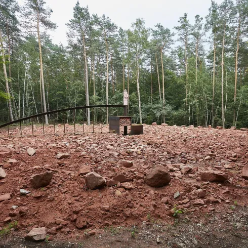 Eine kahle Fläche mit vielen roten Steinen, umgeben von Bäumen. In der Mitte steht ein auffälliges Objekt mit einer gebogenen Struktur.