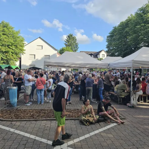 Eine Menschenmenge versammelt sich auf einem Fest mit mehreren weißen Zelten. Im Hintergrund sind grüne Bäume und ein blauer Himmel zu sehen.