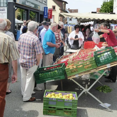 Ein Marktplatz mit vielen Menschen, die frische Produkte kaufen. Obst und Gemüse sind in Kisten arrangiert, während Verkäufer mit den Kunden interagieren.