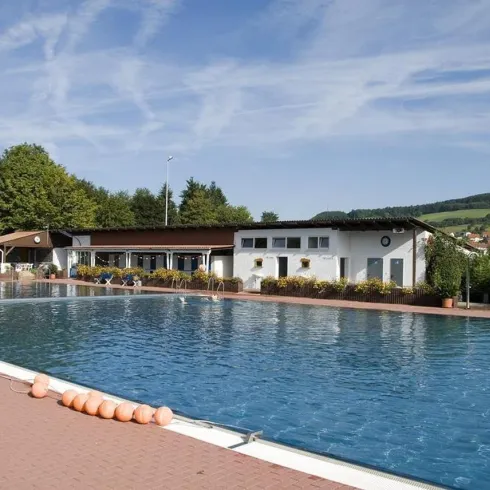 Ein Freibad mit klarem Wasser und einer gemütlichen Umgebung. Im Hintergrund sind Bäume und ein Gebäude zu sehen.