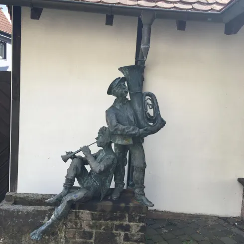 Eine bronzene Statue zweier Musiker steht vor einer weißen Wand. Einer spielt auf einer Flöte, während der andere eine Tuba hält.