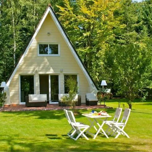 Ein gemütliches Ferienhaus mit einem spitzen Dach steht in einem grünen Garten. Vor dem Haus befindet sich ein Tisch mit Stühlen, umgeben von Bäumen.