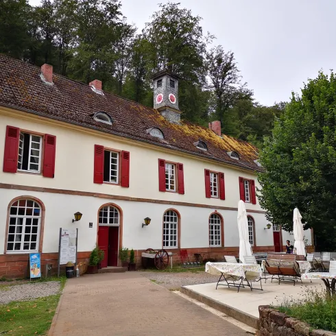 Ein historisches Gebäude mit roten Fensterläden und einem Turm. Der Gartenbereich ist mit Tischen und Stühlen ausgestattet, umgeben von Bäumen.