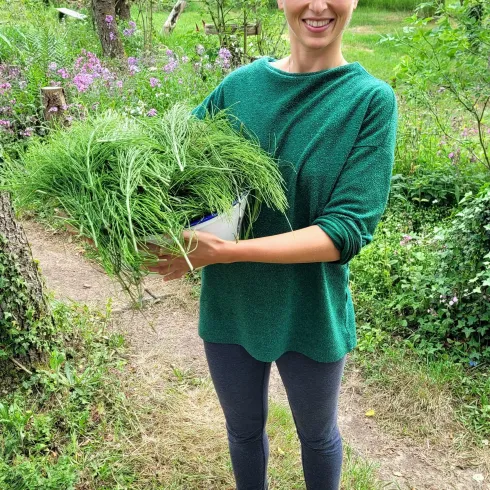 Eine Person steht barfuß im Freien und hält frische Kräuter in der Hand. Im Hintergrund sind grüne Pflanzen und Blumen zu sehen.