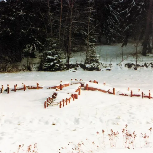 Eine verschneite Landschaft mit skulpturalen Formen aus Holz. Umgeben von Bäumen und einer ruhigen Winteratmosphäre.