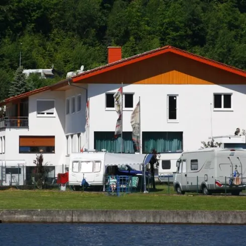 Ein großes teilweises Wohngebäude mit orangefarbenem Dach. Vor dem Gebäude stehen mehrere Wohnmobile und es gibt einen See in der Nähe.