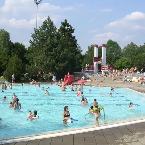 Ein lebhaftes Schwimmbad mit vielen Menschen, die im Wasser spielen. Im Hintergrund sind Rutschen und grüne Bäume zu sehen.