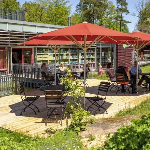 Eine gemütliche Terrasse mit roten Sonnenschirmen und Holzstühlen. Menschen sitzen entspannt und genießen die sonnige Umgebung.