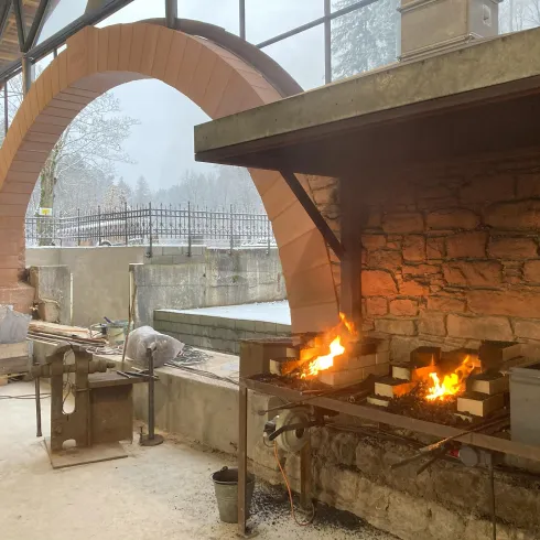 Eine Schmiede mit offenen Feuerstellen und Werkzeugen. Im Hintergrund ist ein großer Bogen und eine schneebedeckte Landschaft zu sehen.