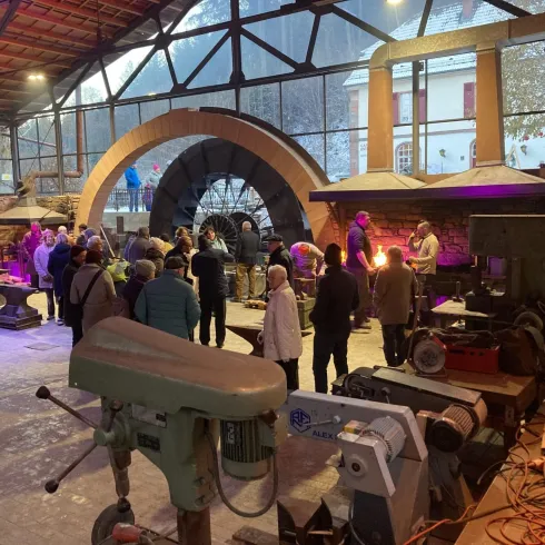 Eine Gruppe von Menschen steht in einer industriellen Umgebung mit Werkstattausrüstung. Im Hintergrund sind große Bögen und Lichtinstallationen zu sehen.